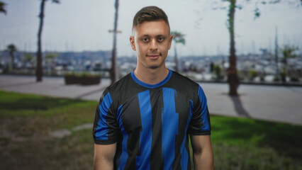 Naklejka premium Man smiling with visible face and tilted head wearing blue soccer jersey on street promenade by marina boats, hands relaxed at sides; pride team spirit.