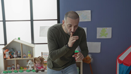 Fototapeta premium Man with hands gesturing and finger to lips for silence in a kindergarten playroom in a school building; patience classroom guidance.