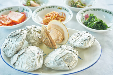 A close up macro of traditional Asian seafood dining featuring fresh hard clams and mixed grain rice. Foil wrapped steamed shells and vibrant side dishes highlight authentic regional coastal gastronom © 진영김
