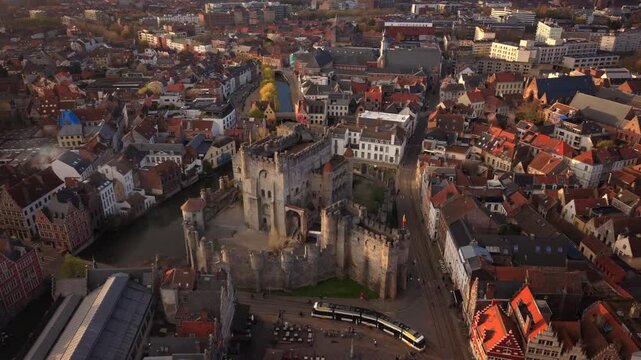 Cinematic aerial Ghent Belgium showcases the majestic Gravensteen Castle surrounded by a moat with traditional Flemish architecture and red-roofed buildings in warm golden hour light.