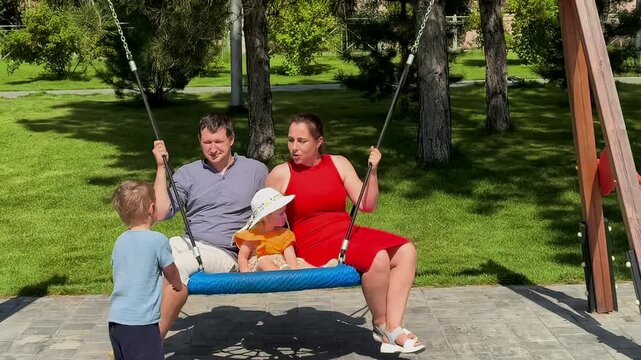 Boy pushes a playground swing with his parents and little sister nearby, family recreation, childhood play, joyful outdoor bonding.