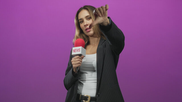 Woman reporter holding red microphone labeled news points finger to camera in studio purple backdrop wearing blazer and white top, direct gaze and hand gesture; confidence reporting.