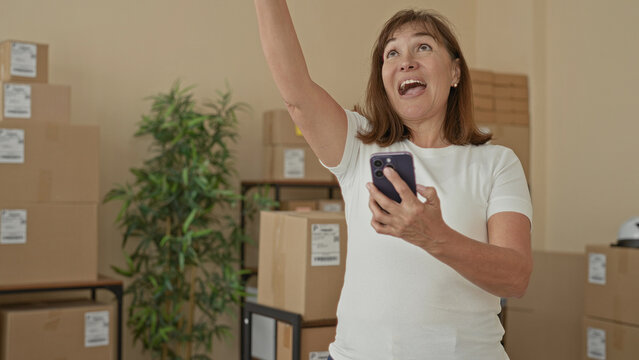 Woman holding smartphone and waving hand in building packed with cardboard boxes and parcels; small business success joy.