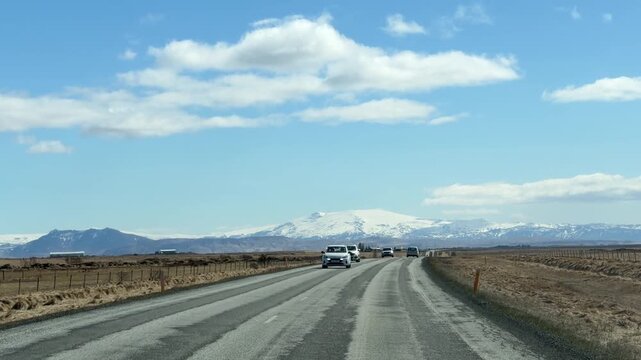 Iceland. Snow-capped mountain range visible in the distance along a straight asphalt road, with clear blue sky and sparse vegetation on both sides of the highway