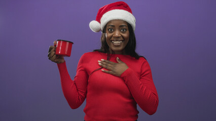 Fototapeta premium Woman in santa hat holds red mug and places hand on chest in studio while smiling and tilting head; joy warmth.