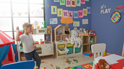 Fototapeta premium Boy throws a small red ball with his right hand inside a colorful children's playroom building scattered with blocks, tent and stuffed toys; playful joy.