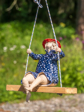 Ceramic Doll On Wooden Swing In Garden Wearing Blue Polka Dot Dress And Red Hat