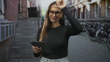 Fototapeta premium Woman holds tablet and makes l sign on forehead on a narrow city street in front of bicycles and brick building; playful teasing.