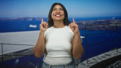 Fototapeta premium Woman pointing index fingers upward on seaside balcony in white sleeveless top and jeans; joy optimism.