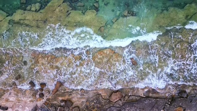 Aerial view of the coastline where waves crash against rocky shores. The scenic seaside showcases nature's beauty with foamy waves and rugged terrain.