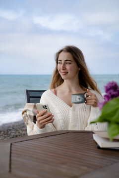 Casual portrait of young woman enjoying coffee and a peaceful moment at a seaside table on a cloudy day, coastal lifestyle and relaxation concept.