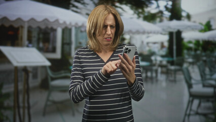 Fototapeta premium Woman tapping smartphone with visible hands and furrowed expression at a street cafe terrace, holding phone while scrolling through messages; everyday tech frustration.