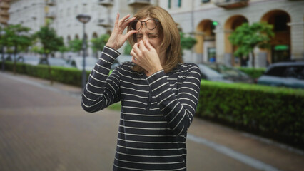 Fototapeta premium Woman in a striped longsleeve top adjusts glasses and repeatedly rubs eyes and temple while standing on a city street with hedges and arched buildings; fatigue eye strain.