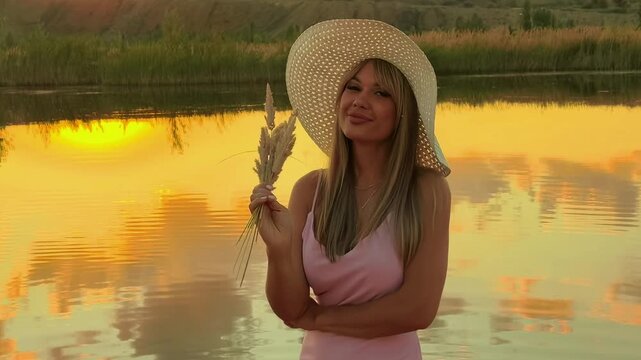 Young woman in a sun hat poses by a lake at sunset, peaceful summer evening, travel lifestyle, scenic outdoor portrait.