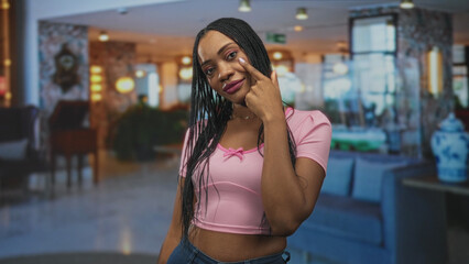 Fototapeta premium Black woman pointing finger to cheek and eye, wearing pink crop top in a building lobby; confidence empowerment poise.