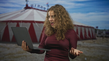 Woman holding laptop, gesturing with open hand and surprised expression in front of a circus tent building  surprise remote work. © Krakenimages.com