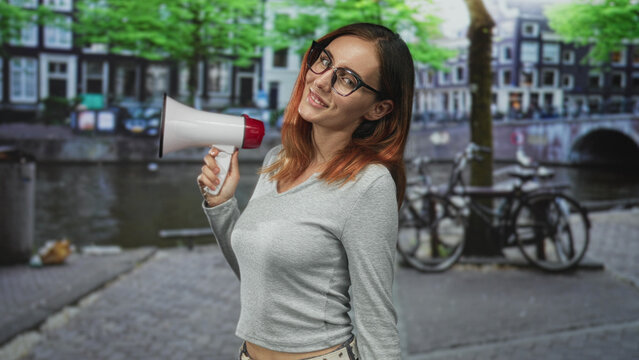 Woman holding megaphone with hand and smiling slightly, standing on street by canal and parked bicycles; voice protest confidence.