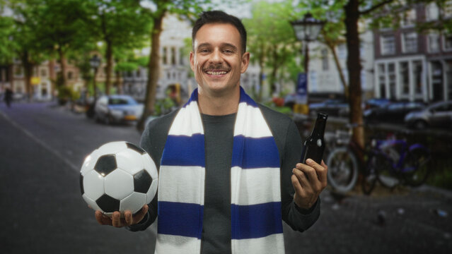 Man holding soccer ball and beer bottle, wearing blue and white scarf, looking hesitant on street; ambivalence support team spirit.