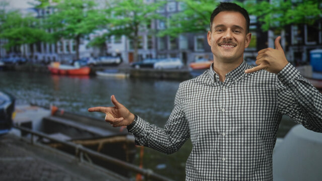 Hispanic man making call-me hand and pointing finger by canal on street, wearing gingham shirt with visible hands and moustache; playful confidence.