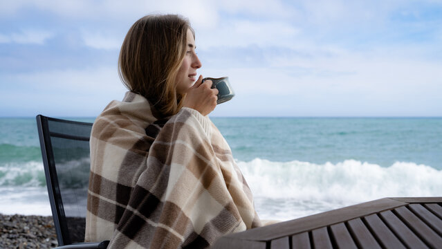 Woman wrapped in a blanket enjoying a warm drink beside the sea on a cool cloudy day, cozy lifestyle and relaxation concept.