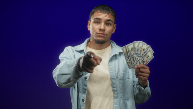 Young man holding fanned us dollars, pointing finger at camera and touching temple in studio; confidence ambition reward.