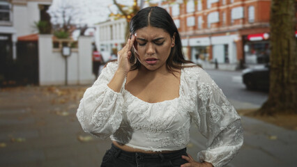 Fototapeta premium Plus size hispanic woman pressing hand to temple for headache on urban street near brick building wearing white embroidered blouse and jeans; discomfort stress.
