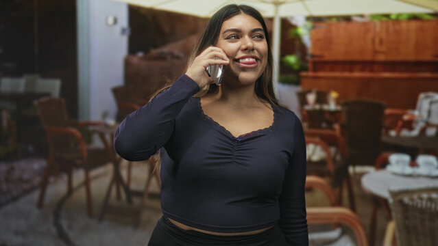 Woman holding phone to ear, smiling with closed eyes and grinning at street cafe; contentment casual chat.