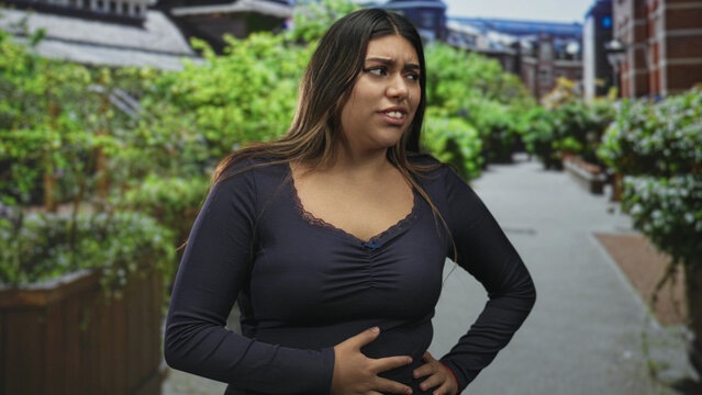 Woman clutching stomach with both hands, eyes closed in a pained grimace, standing on a street lined with planters and brick buildings and a sidewalk; pain nausea.