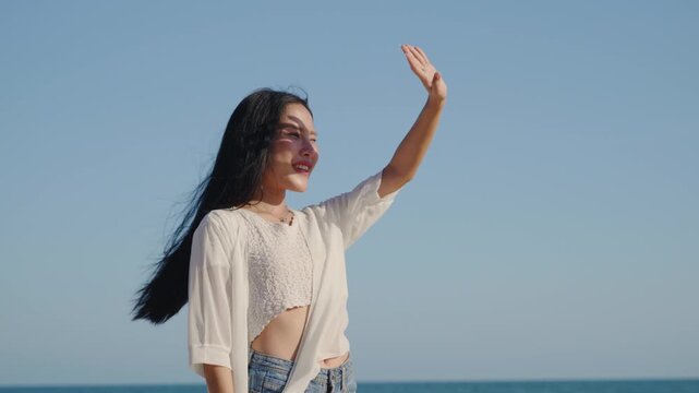 Woman enjoying summer vacation on beach, shielding eyes from sun under clear blue sky, smiling happy Asian female tourist wearing casual white clothes stands near calm ocean.