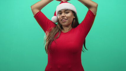 Fototapeta premium Woman plus size hispanic raises arms holding santa hat above head in studio with green backdrop wearing red dress and smiling; holiday festive playful joy.