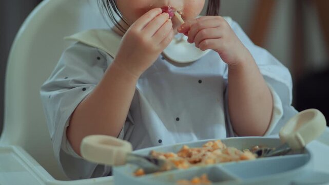 Asian toddler eating spaghetti with hands while sitting in high chair, messy mealtime and independent self feeding at home