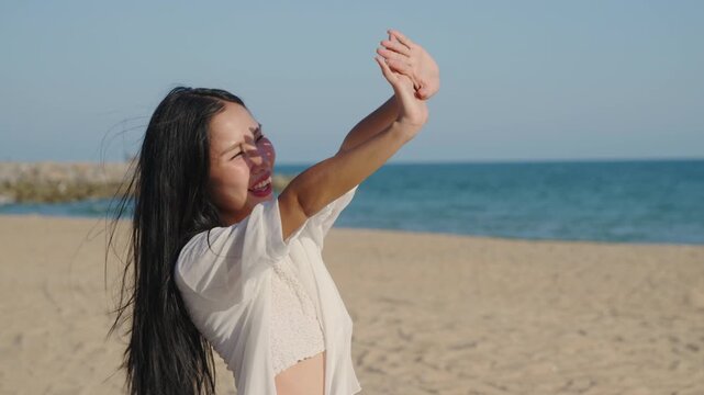Woman enjoying summer vacation on beach shielding eyes from sun, smiling happy asian girl wearing white shirt raising hands to block bright sunshine with blue ocean in background.