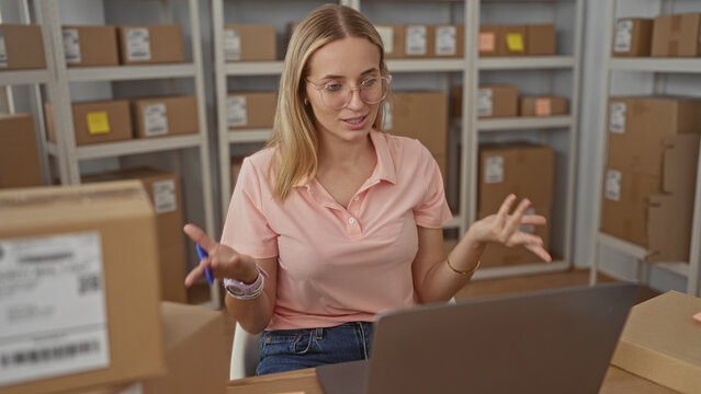 Woman writing sticky note by laptop among stacked cardboard boxes in a packing building; focused productivity.
