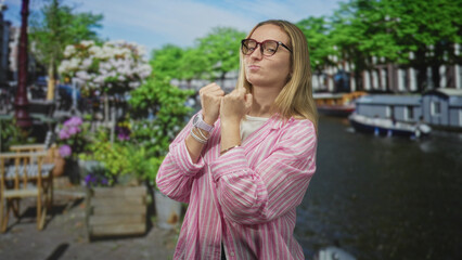 Fototapeta premium Blonde woman with clenched fists near chin on street by canal, wearing glasses and a pink striped shirt; playful confidence.