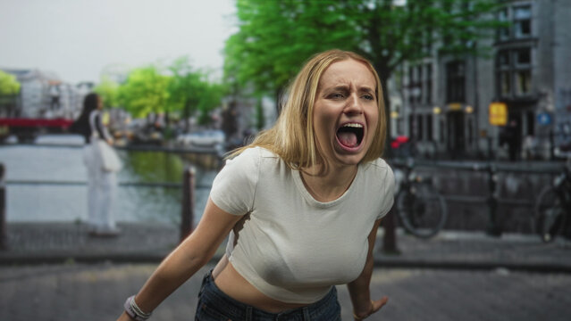 Woman shouting with open mouth and visible midriff in a cropped white tshirt on a city street by a canal and bridge; frustration release.