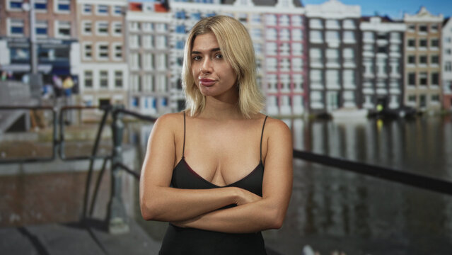 Woman with visible cleavage and crossed arms standing before canal building, neutral gaze and folded posture; defiance.
