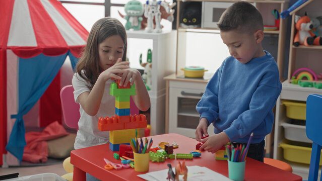 Girl and boy stack colorful plastic blocks with hands at a red table inside a building playroom; concentration cooperation play.