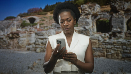 Fototapeta premium Woman holding smartphone, tapping with hands while looking down at the screen amid weathered stone ruins building and gravel foreground; quiet contemplation.
