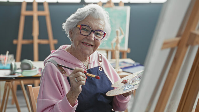 Senior woman holds brush and palette while painting on canvas in studio; creative focus inspiration serenity.