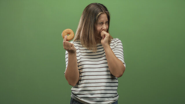Young hispanic blonde woman holding glazed donut with hand to mouth and gagging against green studio backdrop; disgust regret.
