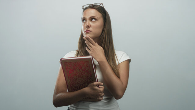 Woman holding ornate red hardcover book, finger pressed to chin in studio with glasses on head and a slight pensive expression; thoughtful curiosity reflection.