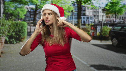 Fototapeta premium Woman wearing santa hat on street holding smartphone to ear and pressing finger to ear, mouth open as if arguing; holiday frustration.