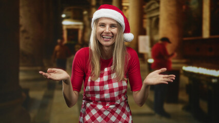 Fototapeta premium Woman wearing santa hat and red checkered apron shows open palms and smiles while shrugging inside a dim building aisle with candles and pillars; holiday cheer.