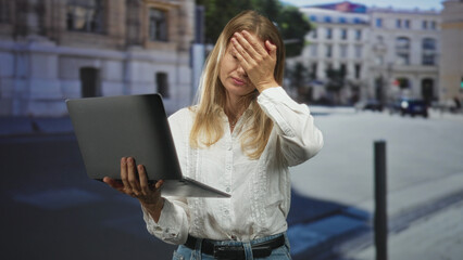 Fototapeta premium Blonde woman holding laptop with hand covering face in city street, facepalm gesture; work frustration.