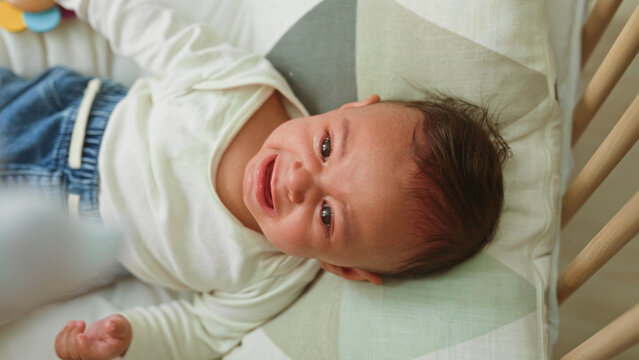 Boy clutching colorful wooden rattle with hand, face scrunched and crying on cradle bed in building; need comfort distress.