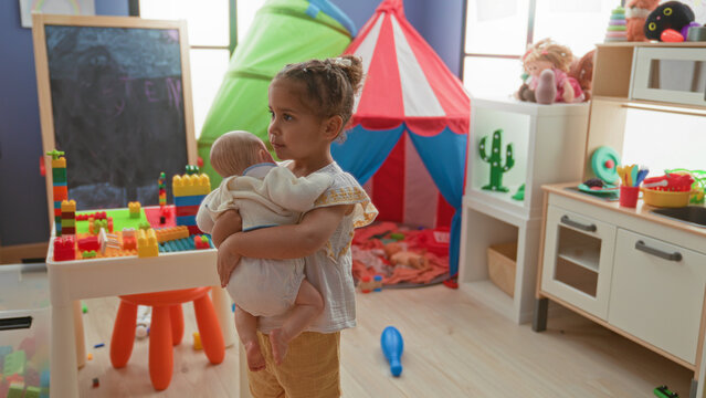 Little hispanic girl cradling baby doll with arms, smiling softly amid building blocks, toy tent and play table in colorful playroom; tenderness.