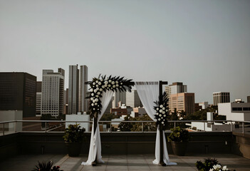 Fototapeta premium Rooftop wedding ceremony arch with white flowers and city skyline background, urban event space and celebration