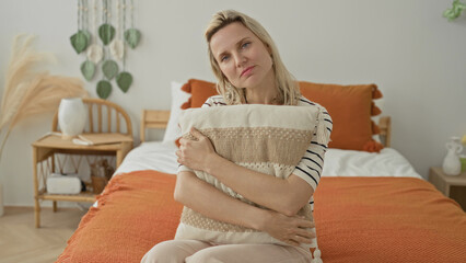 Fototapeta premium Woman hugging pillow on bed inside a building, hands clasped over cushion and orange throw, seated in gentle posture; quiet contemplation solitude.