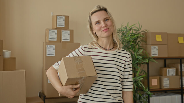 Woman holding a cardboard parcel with one hand and smiling amid stacked shipping boxes in a building; small business pride.