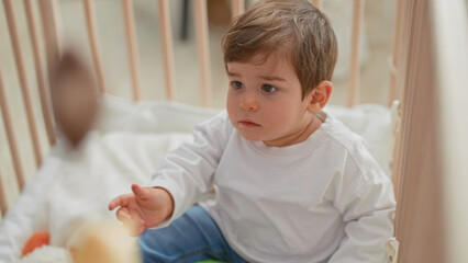 Fototapeta premium Boy reaches with bare hand toward hanging plush mobile above crib in a residential building nursery, face intent and focused on the toy; curiosity.